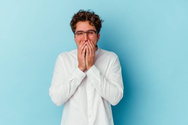 Kafkasyalı genç bir iş adamı mavi arka planda izole edilmiş bir şekilde bir şeye gülüyor, elleriyle ağzını kapatıyor..