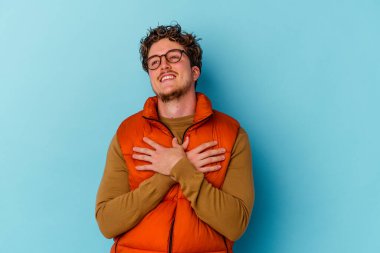 Mavi arka planda izole edilmiş gözlük takan genç beyaz adam, dost bir ifadeye sahip, avuçlarını göğsüne bastırıyor. Aşk konsepti.