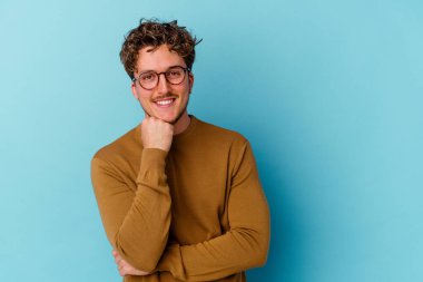 Mavi arka planda izole edilmiş gözlük takan genç beyaz adam mutlu ve kendinden emin gülümsüyor, elleriyle çenesine dokunuyor..