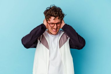 Mavi arka planda gözlük takan genç beyaz adam kulaklarını elleriyle kapatıyor..