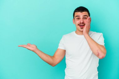 Mavi arka planda izole edilmiş genç beyaz bir adam avucunun içinde fotokopi alanı tutuyor, el üstünde tutuyor. Şaşırmış ve mutlu..