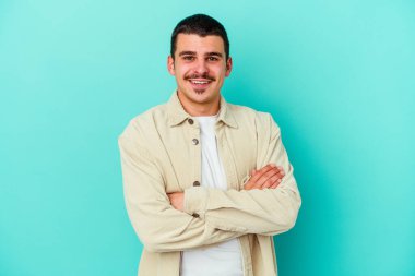 Mavi arka planda kendini güvende hisseden, kararlılıkla kollarını kavuşturan genç beyaz bir adam..