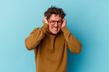 Mavi arka planda gözlük takan genç beyaz adam kulaklarını elleriyle kapatıyor..