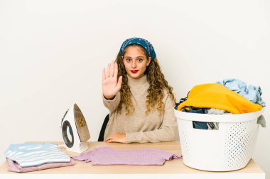 Kafkasyalı genç bir kadın kıyafetlerini ütülüyor elini uzatmış, dur işareti gösteriyor ve seni engelliyor..