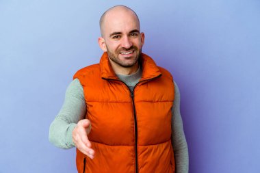 Mor arka planda izole edilmiş, genç, beyaz, kel bir adam. Kameraya doğru elini uzatarak selam veriyor..