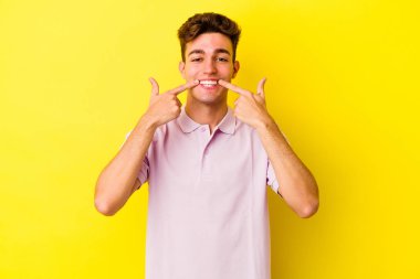 Sarı arka planda izole edilmiş genç beyaz adam gülümsüyor, parmaklarını ağzına doğrultuyor..