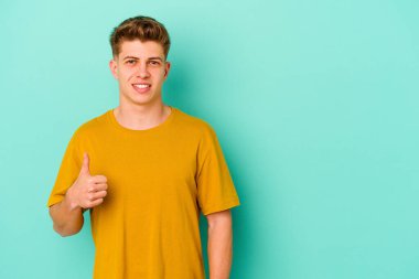 Mavi arka planda izole edilmiş genç beyaz adam gülümsüyor ve başparmağını kaldırıyor.