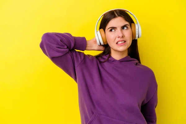 Young caucasian woman listening to music with headphones isolated on pink background touching back of head, thinking and making a choice.