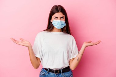 Virüs maskesi takan beyaz bir kadın pembe arka planda izole edilmiş. Kopya alanı tutmak için omuz silkiyor ve kafası karışık..