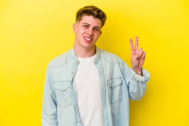 Sarı arka planda izole edilmiş genç beyaz adam zafer işareti gösteriyor ve gülümsüyor..