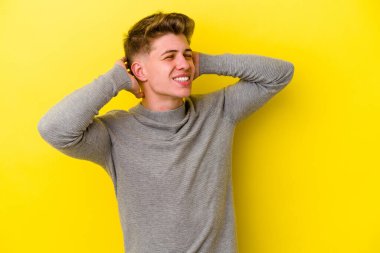 Sarı arka planda izole edilmiş genç beyaz bir adam kendini güvende hissediyor, elleri başının arkasında..