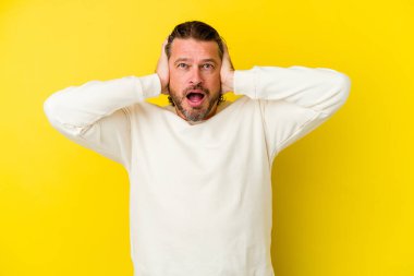 Orta yaşlı, beyaz bir adam sarı arka planda izole edilmiş kulakları kulaklarıyla kapatıyor ve çok ses duymamaya çalışıyor..