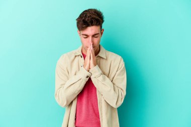 Kafkasyalı genç adam mavi arka planda izole edilmiş dua ediyor, bağlılık gösteriyor, dindar biri ilahi ilham arıyor..