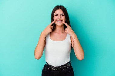 Mavi arka planda izole edilmiş genç beyaz kadın gülümsüyor, parmaklarını ağıza doğrultuyor..