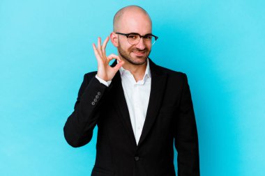 Genç, beyaz, kel bir adam mavi arka planda izole edilmiş bir şekilde bir göz kırpıyor ve elleriyle iyi bir jest yapıyor..