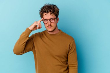 Gözlüklü genç beyaz adam mavi arka planda izole edilmiş. Şakağı parmağıyla işaret ediyor. Düşünüyor, bir göreve odaklanıyor..