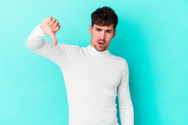 Mavi arka planda izole edilmiş genç beyaz bir adam baş parmağını aşağı gösteriyor ve hoşlanmadığını ifade ediyor..
