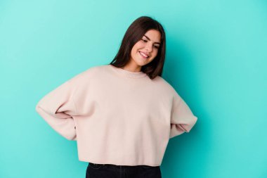 Kafkasyalı genç bir kadın mavi arka planda izole edilmiş. Kendine güvenen, ellerini kalçasında tutan..