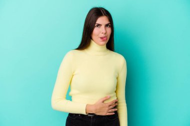 Mavi arka planda izole edilmiş genç beyaz kadın karnına dokunuyor, nazikçe gülümsüyor, yemek yiyor ve tatmin oluyor..