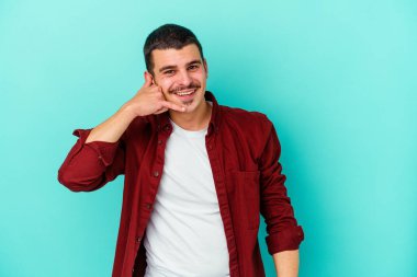 Mavi arka planda izole edilmiş genç beyaz adam parmaklarıyla bir cep telefonu hareketi gösteriyor..