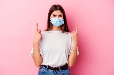 Pembe arka planda virüse karşı maske takan beyaz bir kadın iki ön parmağıyla boş bir alan gösterdiğini gösteriyor..