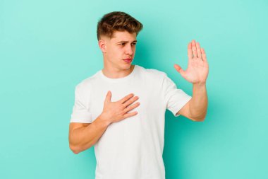 Kafkasyalı genç adam mavi arka planda izole edilmiş yemin ediyor, elini göğsüne koyuyor..