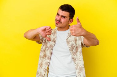 Sarı arka planda izole edilmiş genç beyaz adam baş parmağını kaldırıyor ve baş parmağını aşağı gösteriyor, zor seçim konsepti.