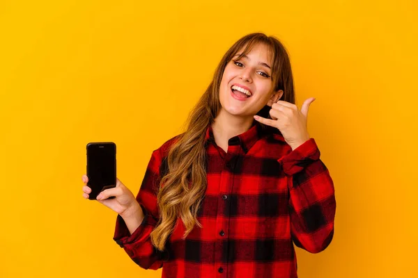 Young caucasian woman holding phone isolated on yellow background showing a mobile phone call gesture with fingers.