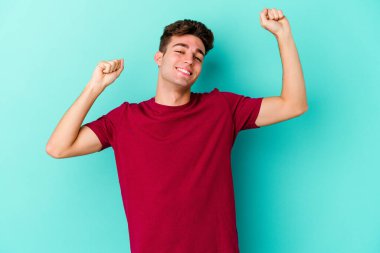 Mavi arka planda izole genç beyaz adam özel bir günü kutluyor, atlıyor ve enerjiyle kollarını kaldırıyor..