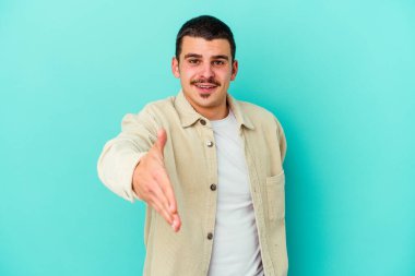 Mavi arka planda izole edilmiş genç beyaz adam selamlama jesti olarak kameraya elini uzatıyor..