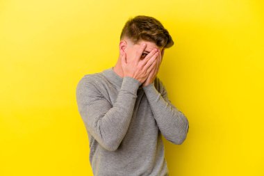 Sarı arka planda izole edilmiş genç beyaz adam parmaklarının arasından korkmuş ve gergin bir şekilde göz kırp..