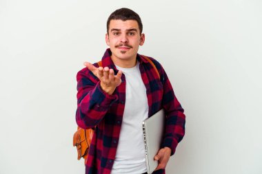 Beyaz arka planda dizüstü bilgisayar tutan beyaz tenli genç bir öğrenci elinde avuç içiyle kameraya sunduğu bir şeyi tutuyor..