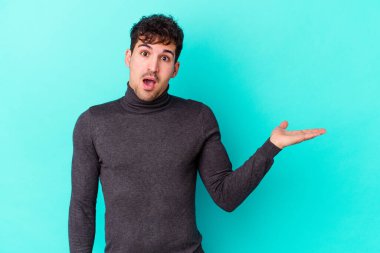 Mavi arka planda izole edilmiş genç beyaz adam, avuç içi fotokopi alanını elinde tutmaktan etkilenmiş..