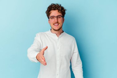 Beyaz tenli genç iş adamı mavi arka planda izole edilmiş. Kameraya doğru elini uzatarak selam veriyor..