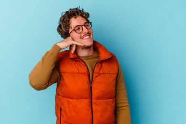 Mavi arka planda izole edilmiş gözlüklü genç beyaz adam parmaklarıyla bir cep telefonu hareketi gösteriyor..