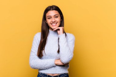 Sarı arka planda izole edilmiş genç Hintli kadın gülümsüyor mutlu ve kendinden emin, elleriyle çenesine dokunuyor..