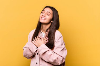 Sarı arka planda izole edilmiş genç Hintli kadın gülüyor ellerini kalbe koyuyor, mutluluk kavramını koruyor..