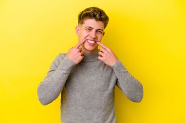 Sarı arka planda izole edilmiş genç beyaz adam gülümsüyor, parmaklarını ağzına doğrultuyor..