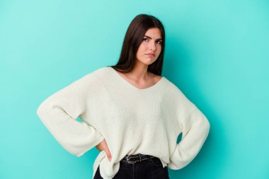 Mavi arka planda izole edilmiş genç beyaz kadın hoşnutsuzca kaşlarını çatmış, kollarını kavuşturmuş..