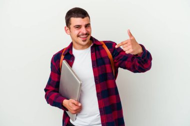 Beyaz arkaplanda dizüstü bilgisayar tutan beyaz tenli genç bir öğrenci gömlek fotokopi alanını işaret ediyor, gururlu ve kendinden emin.