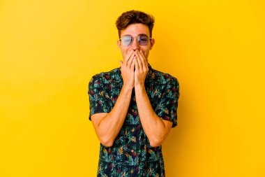 Hawaii gömleği giyen genç beyaz adam sarı arka planda izole edilmiş bir şekilde bir şeye gülüyor, elleriyle ağzını kapatıyor..