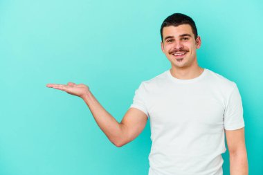 Mavi arka planda izole edilmiş genç beyaz bir adam bir palmiye üzerinde fotokopi alanı ve belinde bir el daha gösteriyor..