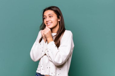 Mavi arka planda izole edilmiş genç Hintli kadın ellerini çenesinin altında tutuyor ve mutlu bir şekilde yan tarafa bakıyor..