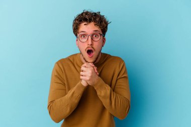 Mavi arka planda izole edilmiş gözlüklü genç beyaz adam şans için dua ediyor, şaşırıyor ve ağzını açıyor..