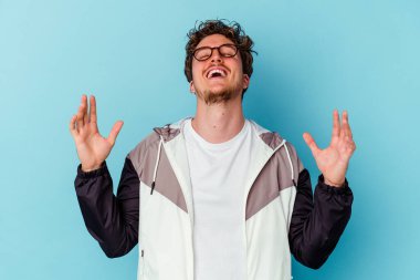 Mavi arka planda izole edilmiş gözlüklü genç beyaz adam yüksek sesle gülüyor ve elini göğsüne koyuyor..