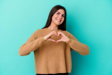 Mavi arka planda izole edilmiş genç beyaz kadın gülümsüyor ve elleriyle kalp şeklini gösteriyor..