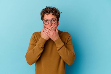 Mavi arka planda gözlüklü genç beyaz adam elleri ile ağzını kapatırken şok oldu..