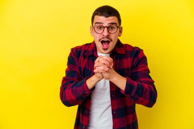 Beyaz tenli genç adam sarı arka planda izole edilmiş şans için dua ediyor, şaşırmış ve ağzını açıp önüne bakıyor..