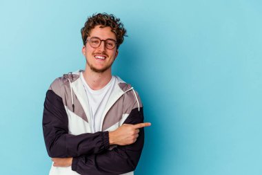 Mavi arka planda gözlüklü genç beyaz adam işaret parmağıyla işaret ederek gülümsüyor..