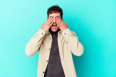 Mavi arka planda soyutlanmış genç beyaz bir adam elleriyle gözlerini kapatıyor, geniş bir gülümsemeyle bir sürpriz bekliyor..